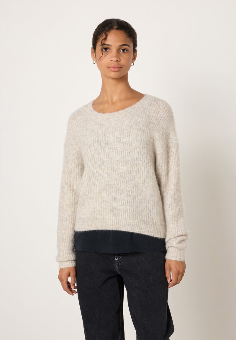 Jauna sieviete ar cirtainiem matiem, valkā zelta apli auskarus, mīkstu bēša trikotāžas džemperi pār melnu kreklu un tumšas džinsa bikses pret vienkāršu fonu.