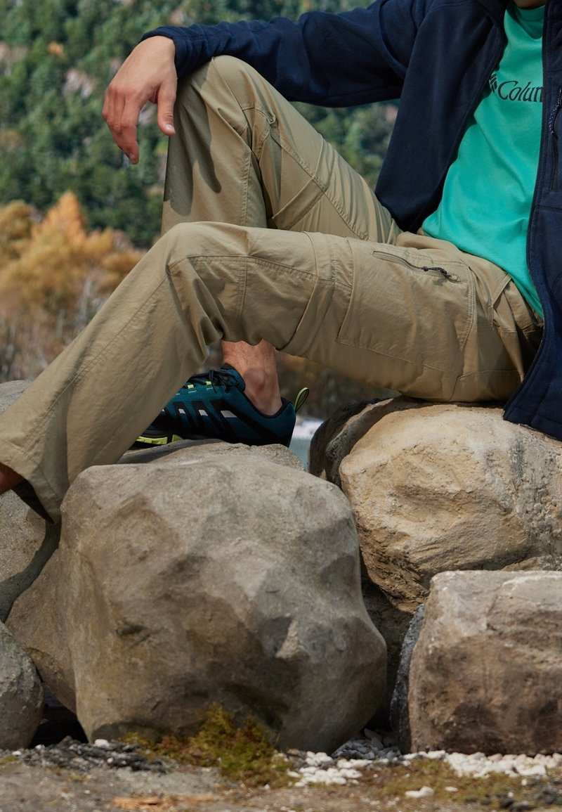 Camicia di tessuto verde, giacca blu navy e pantaloni cargo kaki. Scarpe da trekking blu con dettagli verdi. Persona seduta su grosse rocce all'aperto.
