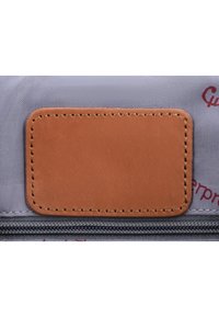 Patch en cuir marron cousu sur un tissu gris au-dessus d'une fermeture éclair noire, partie d'un sac ou d'un étui avec un texte rouge imprimé partiellement visible.