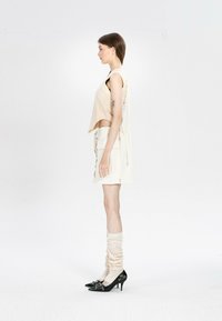 Mujer de perfil vestida con un top beige sin mangas, falda crema con lazos laterales, calentadores de piernas y tacones negros puntiagudos sobre un fondo liso.