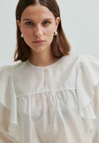 Młoda kobieta z brązowymi włosami do ramion, nosząca srebrne kolczyki w kształcie koła oraz białą bluzkę z falbaniastymi rękawami, która patrzy bezpośrednio w obiektyw.