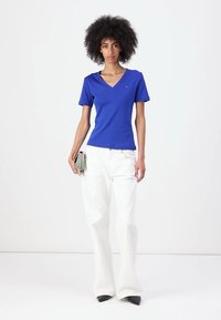 Blauwe katoenen v-hals T-shirt met korte mouwen, gecombineerd met witte flare-broeken en zwarte puntige hakken. Model houdt een kleine handtas vast.