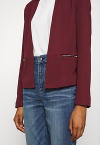 Personne portant un jean bleu, une chemise blanche et une veste rouge foncé avec des poches zippées visibles, se tenant devant un fond uni.