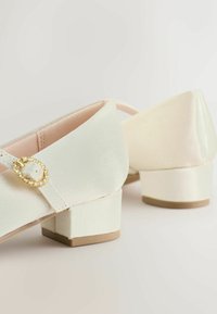 Pantofi din satin alb cu un toc mic bloc, având o cataramă circulară aurie cu strasuri pe o baretă. Textură netedă, design minimalist.