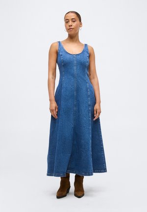 Woman standing wearing a sleeveless blue denim maxi dress and brown ankle boots against a plain background.