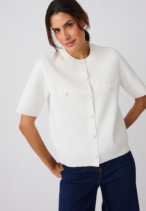 Frau mit dunklen Haaren, die eine weiße kurzärmelige, geknöpfte Strickjacke und dunkelblaue Jeans trägt, steht mit einer Hand in der Tasche.