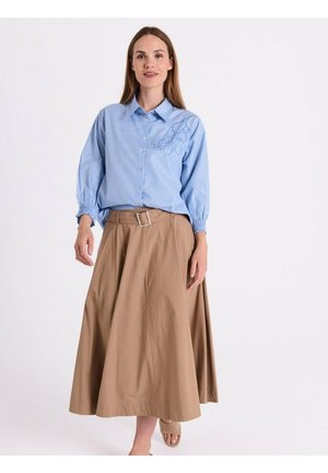 Vrouw draagt een lichtblauw gestreepte blouse met bloemenborduursel en een beige midirok met een riem, staand tegen een effen witte achtergrond.