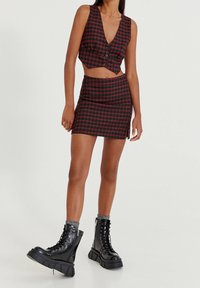 Gilet croisé écossais rouge et noir avec boutons, associé à une mini-jupe ajustée ; porté avec des bottes de combat noires à plateforme. Texture de tissu lisse.