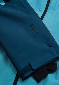Water-resistant jacket in teal and navy. Features a velcro strap and logo. Smooth texture with a streamlined design.