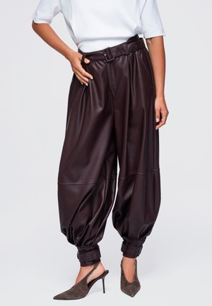 Femme porte un pantalon en cuir marron ample taille haute avec des poignets avec ceinture et un haut blanc à manches courtes, associé à des chaussures slingback grises à talons hauts.