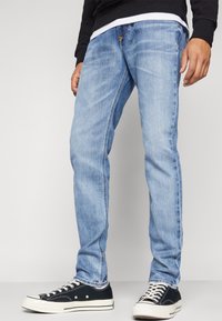 Jeans de mezclilla de color azul claro con un corte ajustado, un desvanecimiento sutil y cinco bolsillos. Combinados con zapatillas de lona negras con suelas blancas.