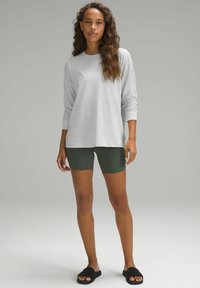 Jasnoszara koszula z długim rękawem zestawiona z ciemnozielonymi szortami sportowymi i czarnymi klapkami. Gładki materiał i luźny krój na całej powierzchni.