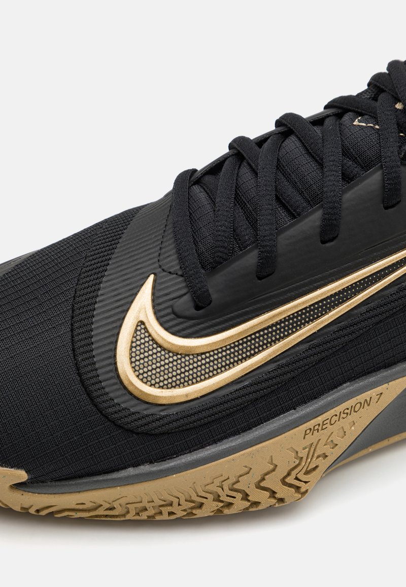 Zapatilla deportiva negra con un upper texturizado, detalles en dorado y una suela de goma con un patrón de zigzag. Presencia de una marca detallada.