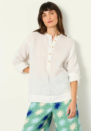 Žena s tmavými vlasmi má na sebe bielu blúzu s dlhými rukávmi a gombíkmi a zelené nohavice s modrými a krémovými slnečnými vzormi, stojaca na jednoduchom pozadí.