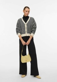 Cardigan grigio e crema con scollatura a V e bottoni frontali, abbinato a pantaloni neri a gamba larga e una borsa gialla. Texture del tessuto liscia.