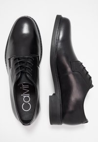 Chaussures de ville en cuir noir avec bout rond, lacets, finition lisse et semelle plate en caoutchouc, étiquetées "Calvin."