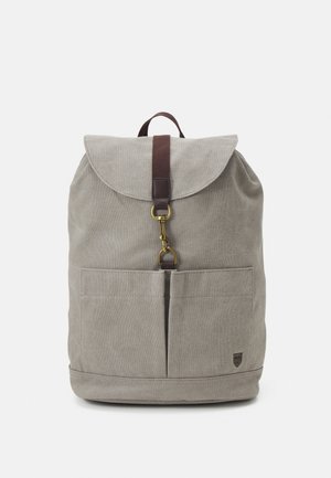 Sac à dos en toile grise avec rabat avant, sangle marron avec boucle dorée, deux poches avant et petit logo noir en bas à droite.