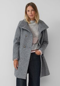 Vrouw met schouderlang blond haar, die zilveren ringoorbellen draagt, een gestructureerde grijze double-breasted jas, een gestreept trui en donkere spijkerbroek.