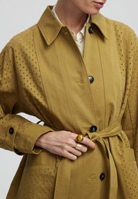 Khaki trenčkot s páskem v pase, zapínáním na knoflíky a perforovanými detaily. Hladká textura s klasickým límcem a bočními kapsami.