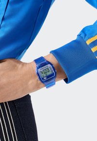 Skaitmeninis laikrodis su skaidria mėlyna plastiko apyranke ir korpusu, turintis skaitmeninį ekraną su baltu tekstu. Nešiojamas ant riešo su mėlyna rankove.