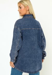 Camicia di jeans blu scuro con una vestibilità rilassata, maniche lunghe e colletto classico. Presenta cuciture chiare e un orlo curvo sul retro.