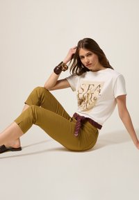 Witte t-shirt met gouden grafische tekst, gecombineerd met mosterdkleurige cropped broek. Accessoires omvatten een gevlochten bordeauxrode riem en een chunky armband.