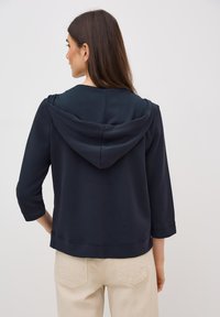 Námořnická modrá mikina s kapucí, volného střihu, tříčtvrtečním rukávem a jemnou texturou. Obsahuje šňůrku na kapuci a jednoduchý design.