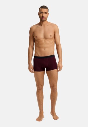 Burgundy boxer shorts with black waistband, fitted design, and soft texture, worn by a standing male model against a neutral background.