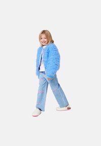 Niebieska puszysta kurtka, graficzna biała koszulka, jasnoniebieskie wytarte dżinsy z różowymi akcentami tekstowymi, białe klapki z różowymi podeszwami. Uśmiechnięte dziecko.