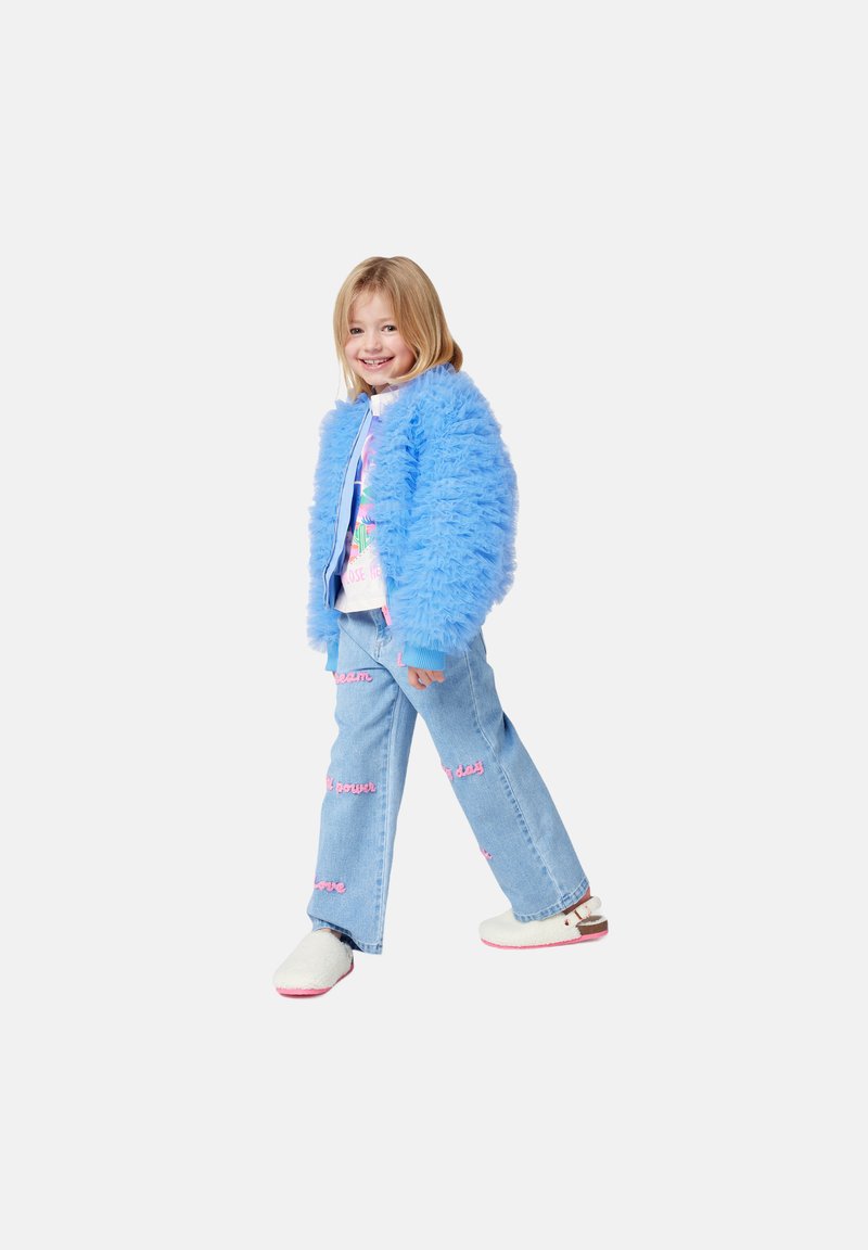 Niebieska puszysta kurtka, graficzna biała koszulka, jasnoniebieskie wytarte dżinsy z różowymi akcentami tekstowymi, białe klapki z różowymi podeszwami. Uśmiechnięte dziecko.