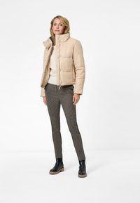 Beige vadderad jacka med hög krage och tryckknappar, bärs över en krämfärgad tröja och mönstrade svart-vita byxor. Svarta ankelstövlar.