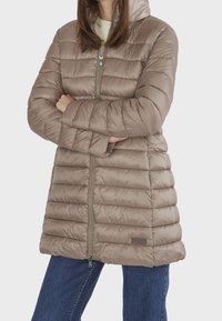Lång vadderad kappa i ljusbrun, med huva, blixtlås fram och horisontellt quiltat mönster. Ärmarna är avpassade med ribbade muddar.