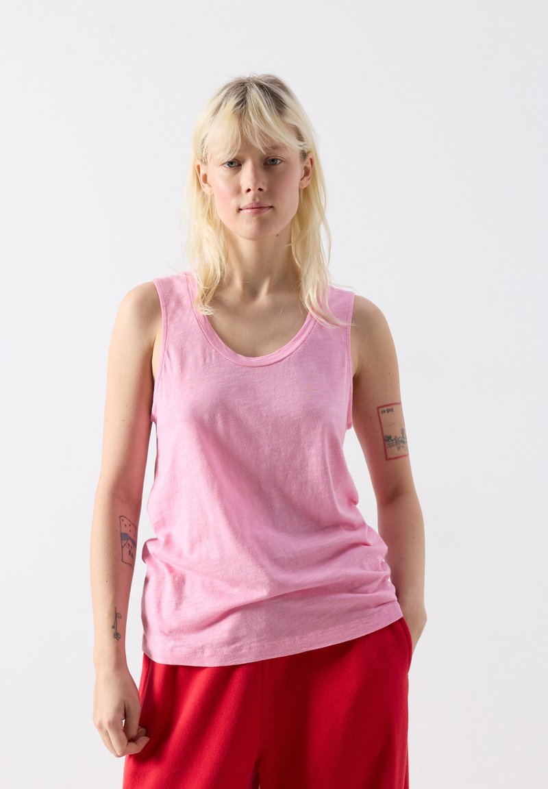 Jauns cilvēks ar blondu matu valkā rozā bezroku kreklu un sarkanas bikses, stāvot pret vienkrāsainu baltu fonu ar tetovējumiem uz rokām.