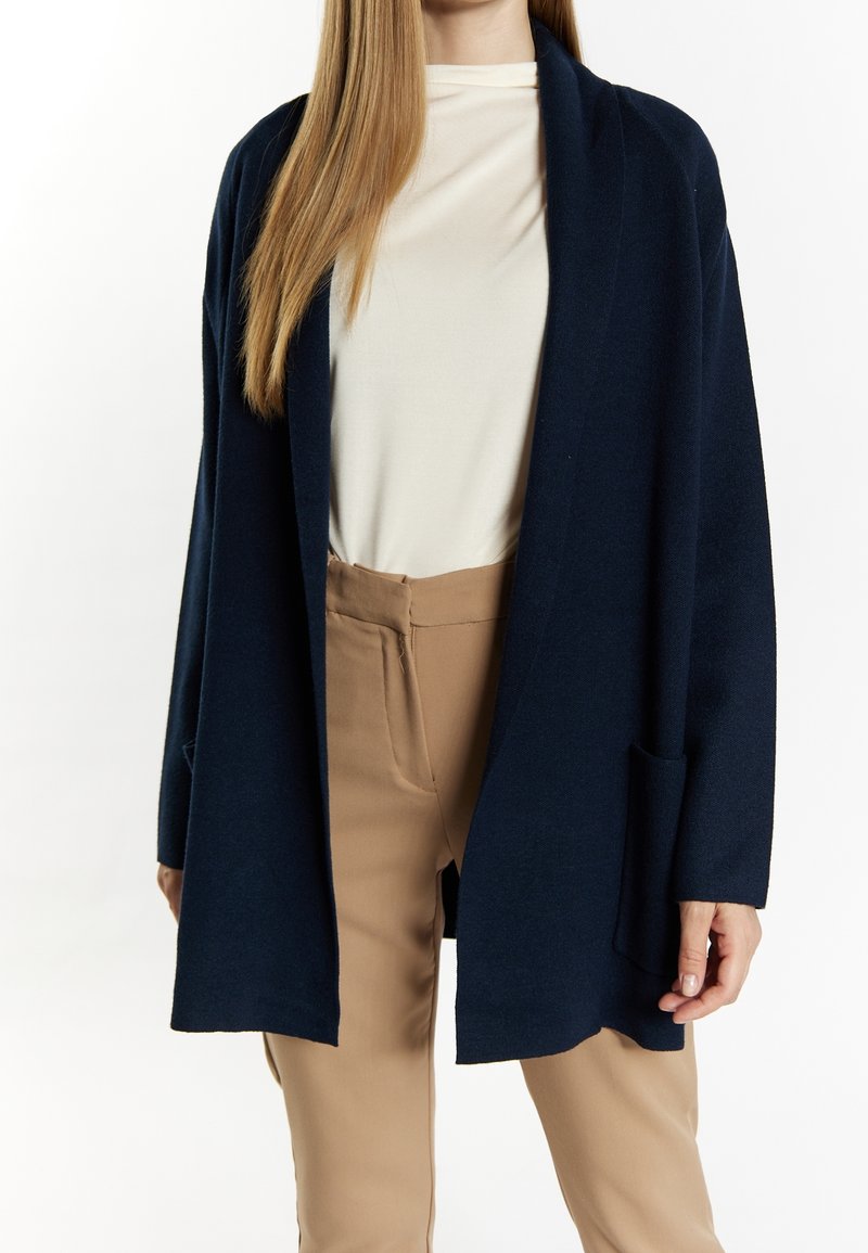 Femme portant un long cardigan bleu marine ouvert, un haut blanc et un pantalon beige, debout sur un fond blanc uni.