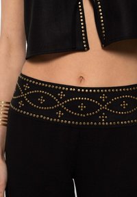 Los pantalones negros presentan una cinturilla ancha adornada con remaches de metal, creando un patrón decorativo. El tejido tiene una textura suave.