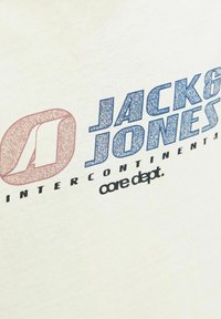 Tkanina boje kreme s grafičkim dizajnom na kojem je ispisano "JACK & JONES" u plavoj boji, crvenim logotipom i tekstom koji kaže "INTER CONTINENTAL core dept."