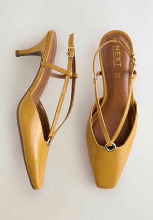 Gule læder slingback-højhælede sko med firkantede tåspidser, med en tynd krydsrem og dekorativt rundt hardware-detalje. Glat tekstur.