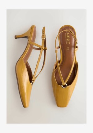Gelbe Slingback-Pumps aus Leder mit quadratischen Zehen, ausgestattet mit einem dünnen Kreuzriemen und dekorativem, rundem Metallakzent. Glatte Textur.