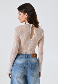 Langärmeliges, transparentes Spitzenshirt in Creme mit floralem Muster und einem Schlüsselloch-Rücken. Getragen mit hochgeschnittenen, hellblauen Jeans.