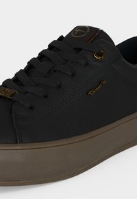 Μαύρο δερμάτινο sneaker με υφή, χοντρή καφέ σόλα, μαύρα κορδόνια, χρυσά κορδόνια και το λογότυπο της μάρκας Tamaris στο πλάι και στη γλώσσα.