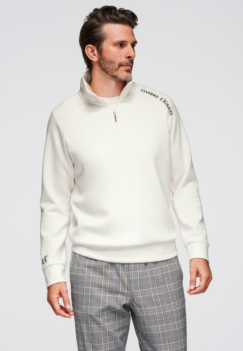 Sudadera blanca con cremallera y cuello alto, con una textura suave y texto negro en el hombro. Combinada con pantalones de cuadros.