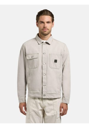 Mężczyzna ubrany w jasnobeżową zapinaną na guziki kurtkę z kieszeniami na piersi oraz dopasowane spodnie typu cargo, stojący na jednolitym białym tle.