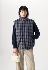 Jūras spēku un pelēka šaha raksta veste ar augstu apkakli, ar rāvējslēdzēju un sānu kabatām, virs tumša garām piedurknēm aug šdaļas, savienota ar gaišām kordūrām.