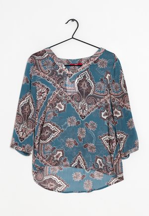Blå bluse med pink, hvide og brune paisley-blomsterprint, trekvartlange ærmer og en rund halsudskæring, hængende på en sort bøjle.