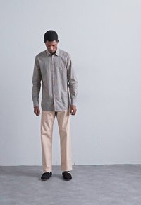 Man in grå rutiga skjortan, beige byxor och svarta skor, som står inomhus mot en enkel ljusgrå vägg.