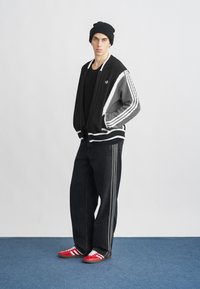 Χοντρό μαύρο και γκρι πλεκτό ζακέτα με λευκές ρίγες, μαύρο t-shirt, φαρδιά μαύρα τζιν και κόκκινα αθλητικά παπούτσια με λευκές ρίγες.