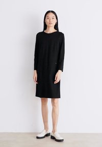 Svart langermet kjole med rund utringing, rett silhuett og knelengde. Bært med hvite loafers og minimal detaljer.