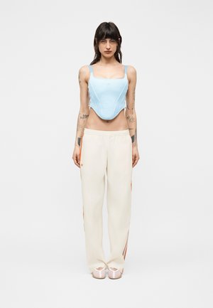 Kobieta o ciemnych włosach ubrana jest w jasnoniebieski gorset, kremowe spodnie dresowe z pomarańczowymi paskami oraz białe trampki, stoi na tle jednolitego tła.