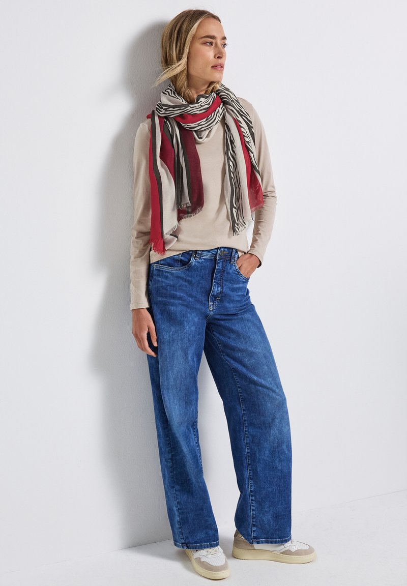 Beige langemouw top gecombineerd met losse blauwe denim jeans. Een rood en zwart gestreepte sjaal hangt om de nek. Lichtgekleurde sneakers maken de look compleet.