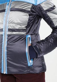 Gros plan sur une personne portant une veste isolante bleu marine, argentée et bleu clair avec une poche zippée, la main dans la poche.